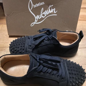 mörkblå sneakers med nitar från Christian Louboutin - Snygga mörkblå sneakers från Christian Louboutin med coola nitar på tån och snörning. Tillverkade i Italien med den ikoniska röda sulan. Perfekta för att ge din outfit en edgy touch.