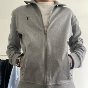 Ralph Lauren tröja - Säljer en stilig grå zip-tröja från Ralph Lauren med broderad logga på bröstet. Tröjan har en dragkedja framtill och långa ärmar. Perfekt för en avslappnad och snygg look. Prisförslag är välkomna!🤩✅