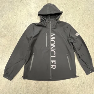 Svart windbreaker från Moncler - Snygg svart vindjacka från Moncler med dragkedja och huva. Jackan har Moncler-logga på ärmen och stort tryck på framsidan. Perfekt för blåsiga dagar.