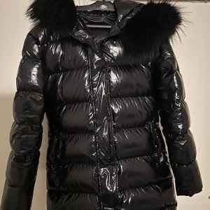 Gloss jacket all black, svart vinterjacka med svart päls - Snygg svart dunjacka med glansigt material och en lyxig pälskrage. Jackan har en dragkedja framtill och långa ärmar. Perfekt för kalla vinterdagar.