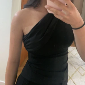 Svart oneshoulder topp - Snygg svart oneshoulder topp med draperad design. Perfekt för en stilren och elegant look. Passar bra till både kjol och byxor. 