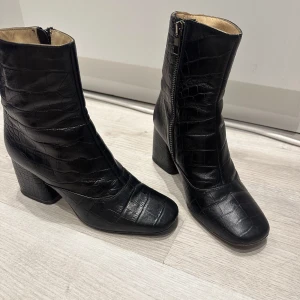 Svarta boots med krokodilmönster från 2NDDAY - Snygga svarta boots i skinn med krokodilmönster och dragkedja på sidan. De har en klack och en spetsig tå, vilket ger dem en elegant look. Perfekta för att lyfta vilken outfit som helst.