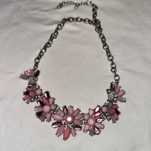 Blommigt halsband i rosa och silver - Charmigt halsband med stora blommor i olika nyanser av rosa och silver. Blommorna är dekorerade med glittriga stenar och sitter på en kedja i silverfärg. Perfekt för att ge en färgklick till din outfit!