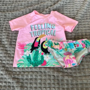 Rosa bikini med tropiskt tryck från H&M - Söt rosa bikini från H&M med tropiskt tryck av tukaner, kaktusar och blommor. Toppen har texten 'Feeling Tropical' och korta ärmar. Bikinitrosorna har volangdetaljer och matchande mönster.