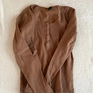 Brun ribbad långärmad topp från Gina Tricot - Säljer en brun ribbad långärmad topp från Gina Tricot. Toppen har en rund halsringning med knappar framtill. Perfekt för en avslappnad stil. 