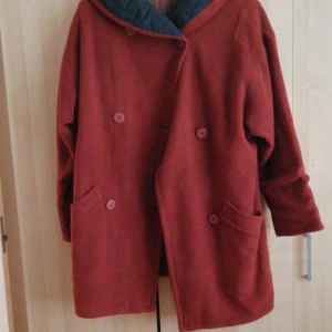 Röd kappa med knappar - Säljer en snygg röd kappa med dubbelknäppning och stora fickor framtill. Den har en unik krage med ett mönstrat foder som ger en cool kontrast. Perfekt för höst och vinter!