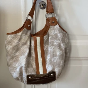 Beige och brun axelväska från Sembonia - Snygg beige axelväska från Sembonia med bruna detaljer och ett mönster av logotyper. Väskan har en praktisk dragkedja på baksidan och två robusta handtag. Perfekt för att bära med sig allt man behöver under dagen.