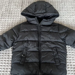 Svart pufferjacka med huva - Säljer en svart pufferjacka med huva och dragkedja. Jackan har långa ärmar och två fickor framtill med knappdetaljer. Perfekt för kyliga dagar.