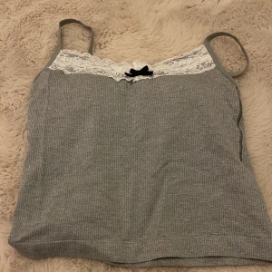 Grå ribbad linnetopp med spetsdetaljer - Säljer en söt grå ribbad linnetopp med vita spetsdetaljer och en liten svart rosett framtill. Perfekt för en avslappnad stil. Toppen har smala axelband och en mjuk känsla. Med inbyggd paddning.