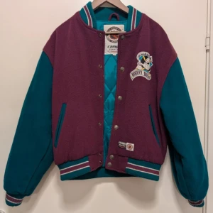 Mighty Ducks jacka från Campri - Säljer en retro Mighty Ducks jacka från Campri i lila och turkos. Jackan har långa ärmar och är dekorerad med broderade logotyper på både fram- och baksidan. Den har knappar framtill och ribbade muddar vid ärmslut och nederkant.
