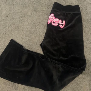 Svarta byxor från Juicy Couture - Snygga svarta byxor från Juicy Couture med rosa broderad logga på baksidan. Byxorna har en bekväm passform med snörning i midjan och är perfekta för en avslappnad stil. De är tillverkade i mjukt material som ger en lyxig känsla.