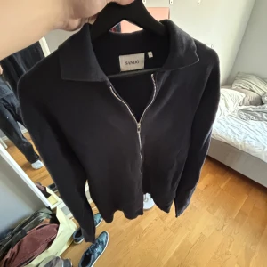 Svart tröja med dragkedja från Sandö - Snygg svart tröja från Sandö med dragkedja framtill. Perfekt för en stilren look. Tröjan har en klassisk krage och är tillverkad i ett mjukt material.