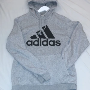 Grå hoodie från Adidas - Säljer en klassisk grå hoodie från Adidas med stor svart logga på framsidan. Tröjan har en bekväm passform med långa ärmar och en praktisk magficka. Perfekt för en avslappnad stil. Hör av er vid frågor.