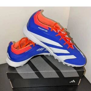 Adidas Predator fotbollsskor - Snygga Adidas Predator fotbollsskor i blått med vita och röda detaljer. Skorna har en modern design med snörning och är perfekta för konstgräs. De har en slitstark sula med bra grepp.