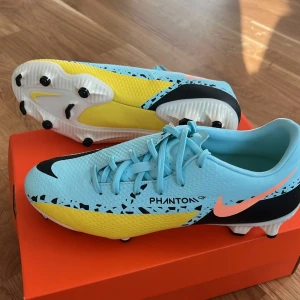 Nike Phantom GT fotbollsskor - Helt nya, bara testade! Snygga ljusblå Nike Phantom GT fotbollsskor med svarta och gula detaljer. Skorna har snörning och en iögonfallande design med en stor Nike-logga på sidan. Perfekta för fotbollsplanen med sina dobbar för bra grepp.