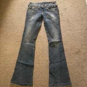 Blå bootcut jeans från True Religion - Snygga blå bootcut jeans från True Religion med ikoniska bakfickor och dekorativa sömmar. Jeansen har en klassisk femficksdesign och knappar med logotyp. Perfekta för en avslappnad stil med en touch av retro. Storleken i byxorna är 24.