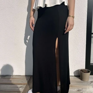 Svart Långkjol från Gina Tricot - Snygg svart lång kjol i superfint skick. Den har en slit och är väldigt skön att ha på varma sommardagar. För referens är tjejen på bilden 168 cm lång.