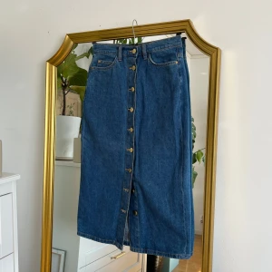 Jeanskjol från Holly & Whyte - Snygg jeanskjol från Holly & Whyte med knappar framtill. Kjolen har en klassisk blå denimfärg och är i en längre modell. Perfekt för en stilren look.
