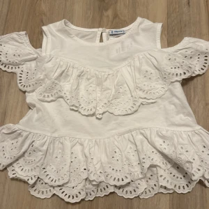 Vit blus från Mayoral - Säljer en söt vit blus från Mayoral med vackra spetsdetaljer och volanger. Blusen har korta ärmar och en knapp i nacken för enkel påklädning. Perfekt för sommarens varma dagar!