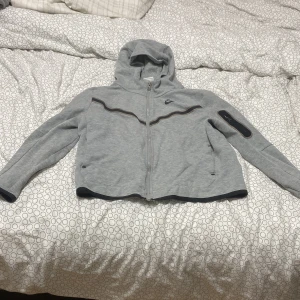 Grå hoodie från Nike - Säljer en grå hoodie från Nike med dragkedja och huva. Jackan har långa ärmar och en liten ficka på ärmen. Perfekt för en sportig look eller en avslappnad dag.