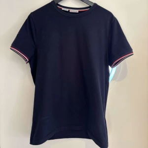 Mörkblå t-shirt från Moncler - Snygg mörkblå t-shirt från Moncler med korta ärmar och stilrena ränder i rött och vitt vid ärmsluten. Perfekt för en casual look med en touch av lyx. Moncler-logga på ärmen ger en exklusiv känsla. L men sitter som M