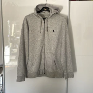 Grå hoodie från Polo Ralph Lauren - Säljer en stilren grå zip hoodie från Polo Ralph Lauren i storlek XL med dragkedja och två fickor framtill. Skriv vid funderingar eller frågor! Lägg ett bud eller köp direkt!