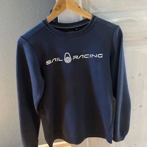 Mörkblå tröja från Sail Racing - Snygg mörkblå tröja från Sail Racing med vit logotyp på bröstet. Tröjan har långa ärmar och en rund halsringning. Perfekt för en stilren och sportig look.