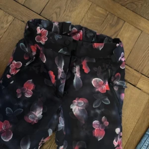 Blommiga täckbyxor från Everest - Snygga täckbyxor från Everest med ett blommigt mönster i rosa på en mörk bakgrund. Byxorna har praktiska dragkedjor och knappar framtill. Perfekta för kyliga dagar.