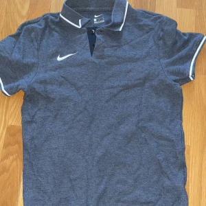 Grå pikétröja från Nike - Säljer en stilren grå pikétröja från Nike med vita detaljer på krage och ärmslut. Perfekt för en sportig look eller avslappnad stil. Tröjan har korta ärmar och en klassisk pikékrage med knappar.