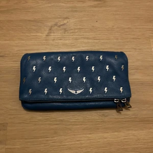 Blå Zadig väska med blixtdetaljer - Snygg blå axelväska från Zadig & Voltaire med coola blixtdetaljer i silver. Väskan har en kedjerem och en praktisk dragkedja på baksidan. Perfekt för att ge din outfit en edgy touch! Finns ett tunt och ett tjockt band till väskan, samt en dust bag! Pris kan fiskuteras!
