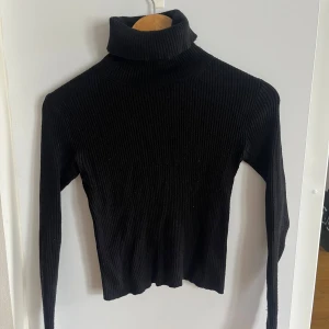 Svart ribbad polotröja från Pull&Bear - Säljer en stilren svart ribbad polotröja från Pull&Bear. Tröjan har en hög krage och långa ärmar, lite croppad. 