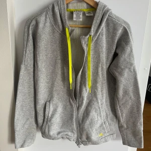 Grå hoodie från Adidas med gula detaljer - Fin grå och knappt använd hoodie från Adidas med dragkedja och gula detaljer. Den har en huva med gula snören och tre gula ränder på ryggen.🌺