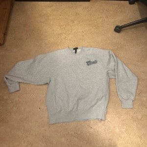 Grå sweatshirt med tryck - Säljer en grå sweatshirt med texten 'Varsity New York' tryckt på ryggen. Tröjan har en klassisk rund hals och långa ärmar. Perfekt för en avslappnad stil.