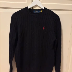 Svart stickad tröja från Polo Ralph Lauren - Säljer en stilren svart stickad tröja från Polo Ralph Lauren med det klassiska röda logotypbroderiet på bröstet. Tröjan har en kabelstickad design och långa ärmar, perfekt för kyligare dagar. Passar både till jeans och chinos för en avslappnad look.