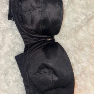 Svart Juicy Couture axelbandslös bh - Snygg svart juicy couture axelbandslös bh med dragkedja framtill. (Mammas från 90 talet) Perfekt för en stilren look. Tillverkad i ett glansigt material som ger en elegant touch.