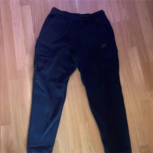 Svarta mjukisbyxor från Nike - Säljer ett par svarta mjukisbyxor från Nike med elastisk midja och dragsko. Byxorna har praktiska cargofickor på sidorna och en diskret Nike-logga. Perfekta för en avslappnad stil.
