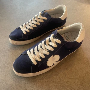 Blå sneakers från The Gallerist - Snygga blå sneakers från The Gallerist med vita detaljer och snörning. Skorna har en unik klöverdesign på sidan och vit sula. Perfekta för en stilren look, Sprillans nya och aldrig använda. Storlek 44. Priset är inte hugget i sten
