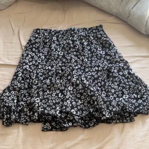 Svart blommig kjol  - Säljer en söt svart kjol med vita blommor från Amisu. Kjolen har en volangdetalj som ger den en superfin look. Perfekt för en somrig dag eller en avslappnad utflykt. Resår i midjan för extra komfort.