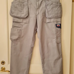 Grå arbetsbyxor från Jobman - Säljer ett par grå arbetsbyxor från Jobman med flera praktiska fickor. Byxorna har en loose passform och är perfekta för arbete med sina slitstarka material och funktionella design. De har också detaljer som knappar och förstärkta sömmar för extra hållbarhet. Midja 96cm. Innemått (insida av ben) 74cm. Hela längden 99cm. Kan hämtas i Vallentuna.