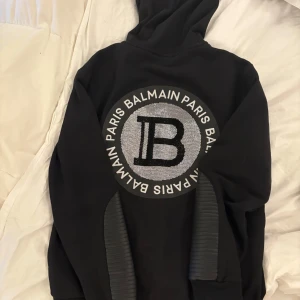 Svart hoodie från Balmain - Säljer en stilren svart hoodie från Balmain med en stor logotyp på ryggen. Hoodien har dragkedja framtill och justerbar huva med snörning. Köpt för 8500kr ca. Den är ganska stor i storleken. Bestäm rimligt pris själv!