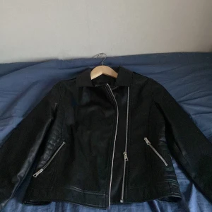 Svart skinnjacka med dragkedja - Snygg svart skinnjacka med asymmetrisk dragkedja och två snedställda fickor med dragkedjor. Jackan har en klassisk krage och quiltade detaljer på sidorna. Perfekt för en cool look!
