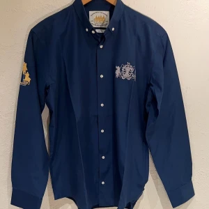 Polo Van Santen Mörkblå skjorta Slim-fit Strl XL(L) - Marinblå skjorta från Polo Van Santen Players Society. Skjortan har broderade detaljer på bröstet och ärmen. Slim fit modell med button-down krage. 100% Bomull I princip oanvända 