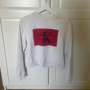 Vit tröja från Calvin Klein Jeans - Säljer en stilren vit tröja från Calvin Klein Jeans med ett stort rött tryck på framsidan. Tröjan har långa ärmar och en rund halsringning. Perfekt för en vardaglig outfit❣️❣️