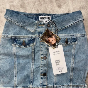 Jeanskjol från H&M x Moschino - Snygg jeanskjol från H&M x Moschino med en unik design som liknar en jeansjacka. Kjolen har knappar framtill och dekorativa fickor med knappar. Perfekt för dig som vill ha en statement piece i garderoben! Kom med bud!