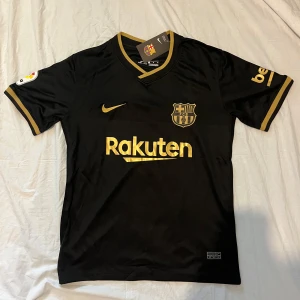 Svart fotbollströja från Nike - Säljer en svart fotbollströja från Nike med guldiga detaljer och logotyper. Tröjan har korta ärmar och V-ringning. Perfekt för fotbollsfans som vill visa sitt stöd med stil.