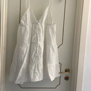 Vit klänning från Zara - Säljer en söt vit klänning från Zara med smala axelband och knytning framtill. Klänningen har en luftig passform och fina spetsdetaljer längs kanterna. Perfekt för varma sommardagar!