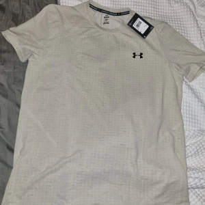 Beige t-shirt från Under Armour - Beige t-shirt från Under Armour med diskret rutmönster och korta ärmar. T-shirten har en klassisk rund hals och är tillverkad i ett lätt och bekvämt material.
