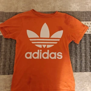 Orange t-shirt från Adidas - Säljer en cool orange t-shirt från Adidas med det klassiska trefoil-logotypen i vitt på framsidan. T-shirten är kortärmad och gjord i mjuk bomull, perfekt för en avslappnad stil. L för barn.