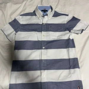Randig skjorta från Tommy Hilfiger - Säljer en stilren randig skjorta från Tommy Hilfiger i blått och vitt. Skjortan har korta ärmar och knappar framtill. Perfekt för en avslappnad men ändå snygg look. 🌀