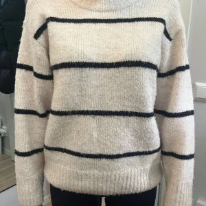 Beige randig stickad tröja från Gina Tricot - Mysig beige stickad tröja från Gina Tricot med svarta ränder. Tröjan har en normal passform och långa ärmar, perfekt för kyligare dagar. Den är tillverkad i ett mjukt material som håller dig varm och bekväm.